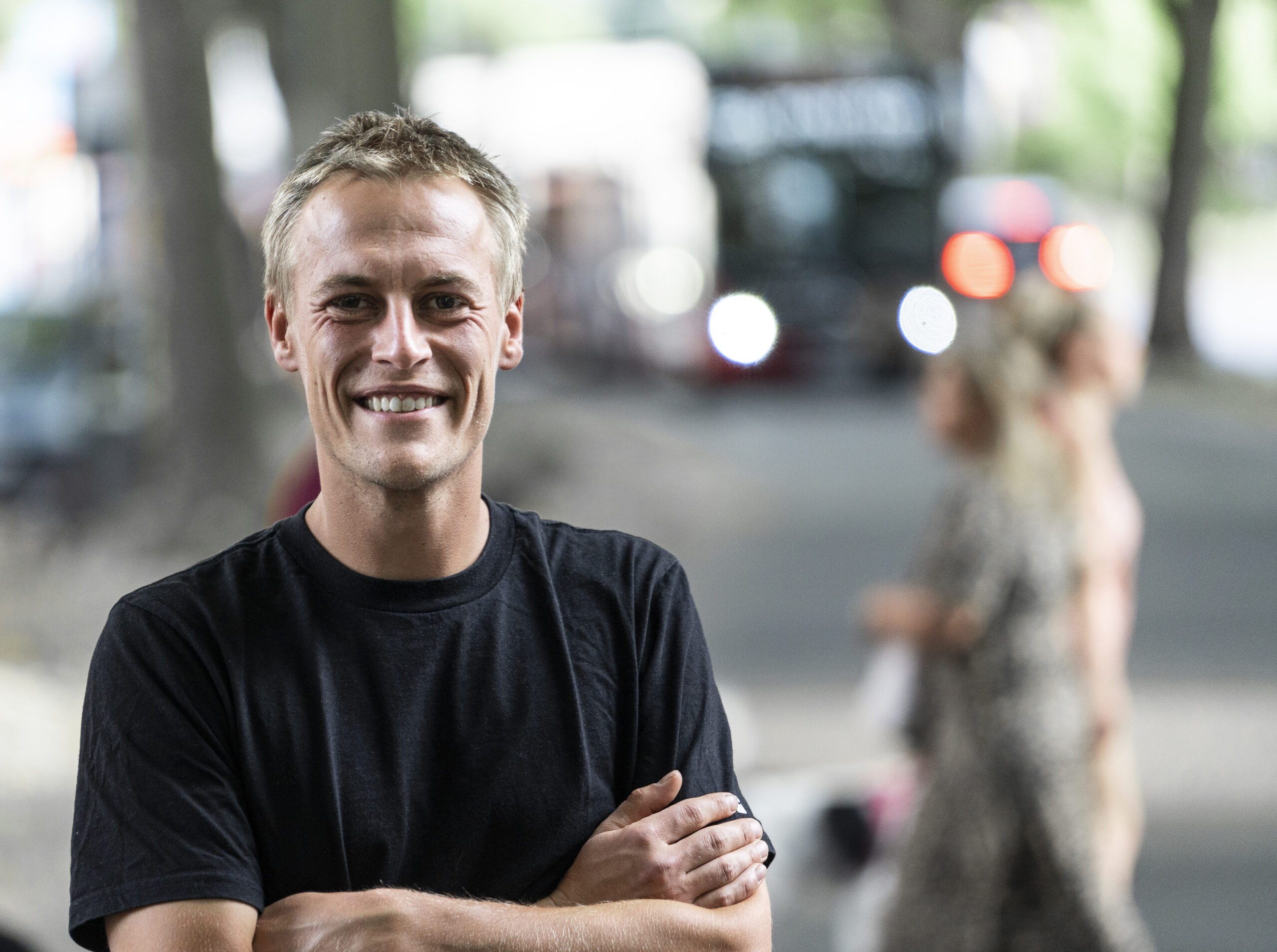 Porträt von Philipp Noack. Philipp hat die Arme verschränkt, trägt ein schwarzes T-Shirt und steht an einer Straßenkreuzung. Im Hintergrund sind verschwommen ein Bus und mehrere Fußgänger*innen zu sehen. 